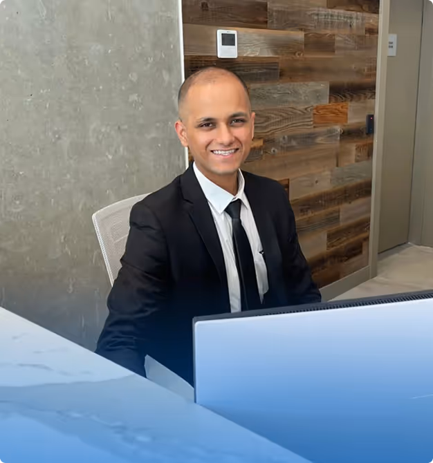 Smiling man in a black suit and tie sitting at a desk with a laptop in a modern office.