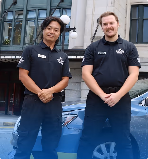 Two security guards in black Blackbird Security uniforms standing in front of a blue car outside a building.