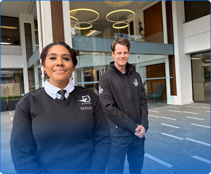 Two security guards in black uniforms standing outside a modern building entrance.