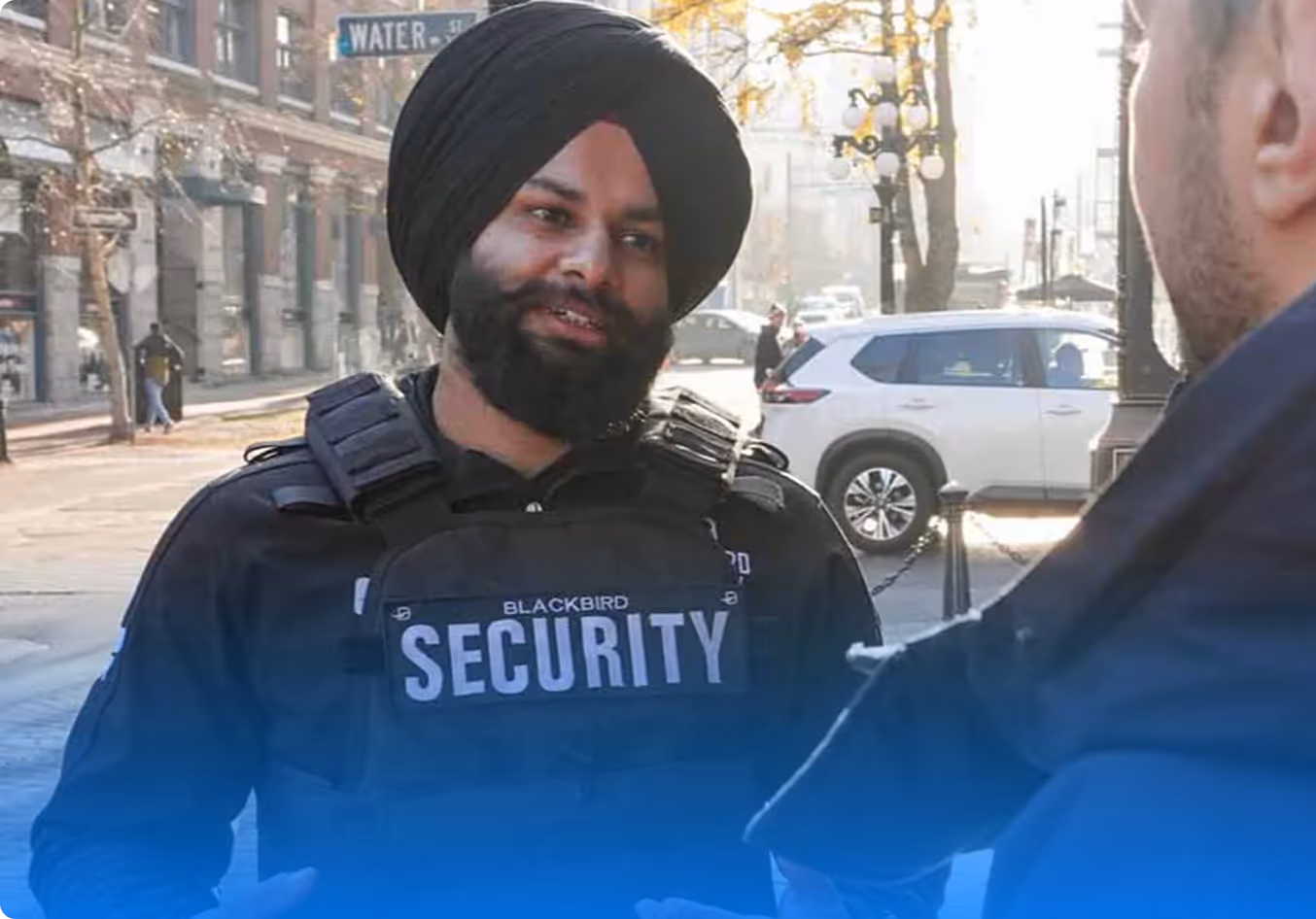 A bearded security guard wearing a black turban and a vest labeled 'Blackbird Security' talking to another person outdoors on a city street.