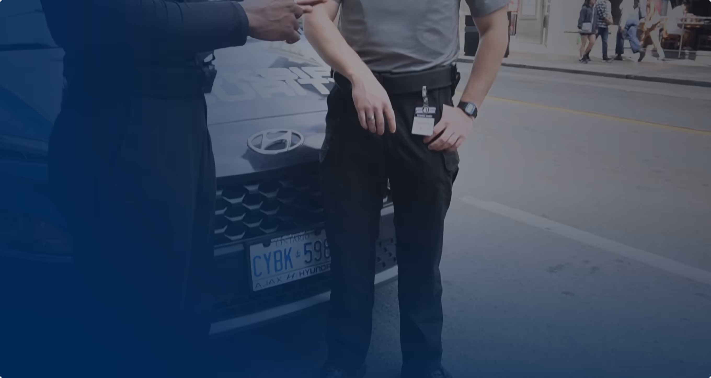 Two security personnel standing in front of a Hyundai vehicle with a visible Ontario license plate on a city street.