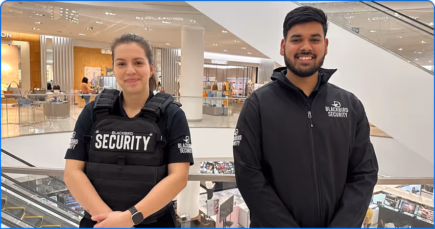 Two security guards wearing Blackbird Security uniforms standing inside a shopping mall near an escalator.