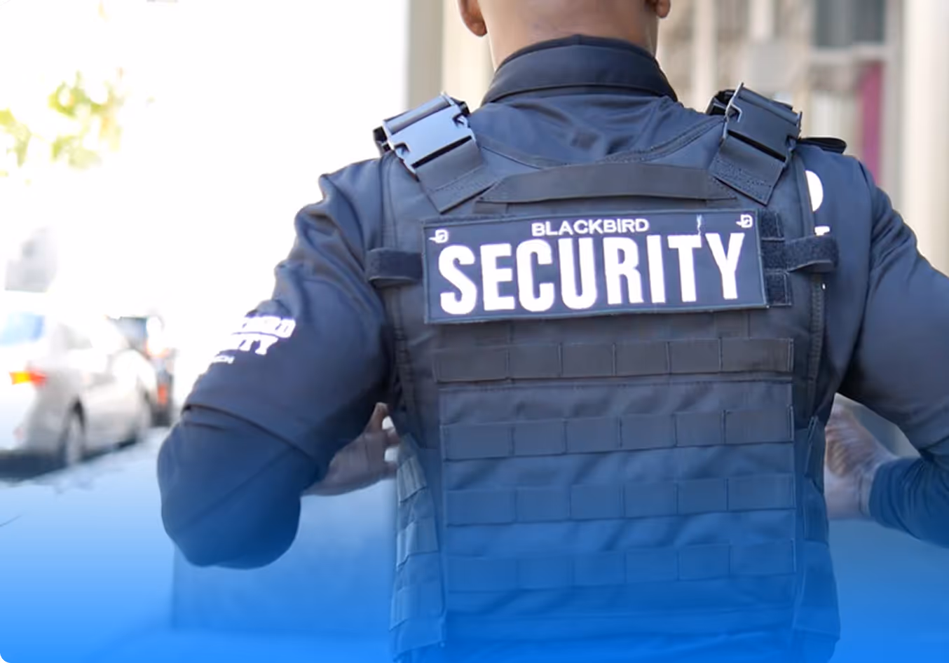 Security officer wearing a Blackbird Security vest seen from behind on a city sidewalk.