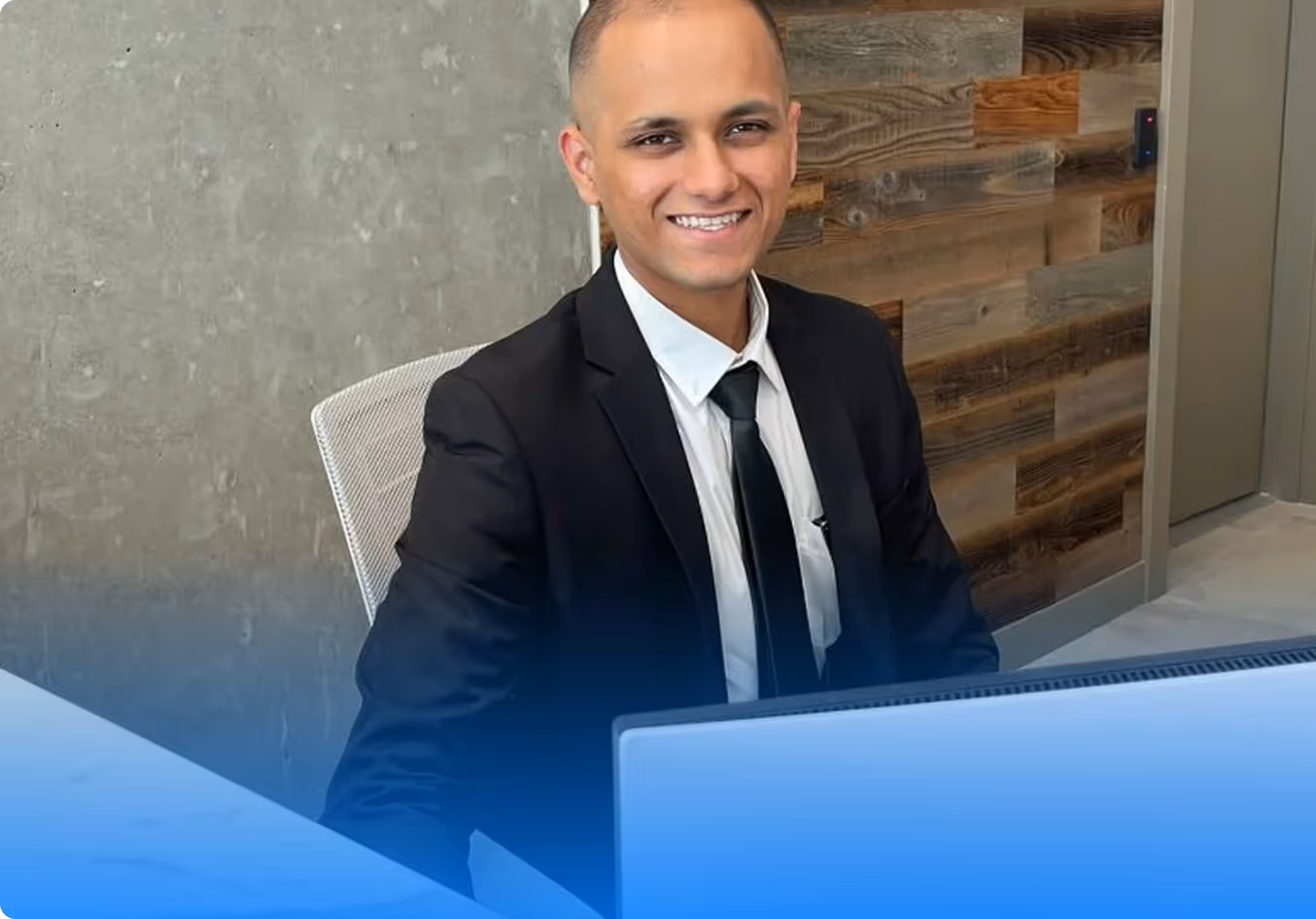 Smiling man in a black suit and tie sitting at a desk with a laptop in a modern office.