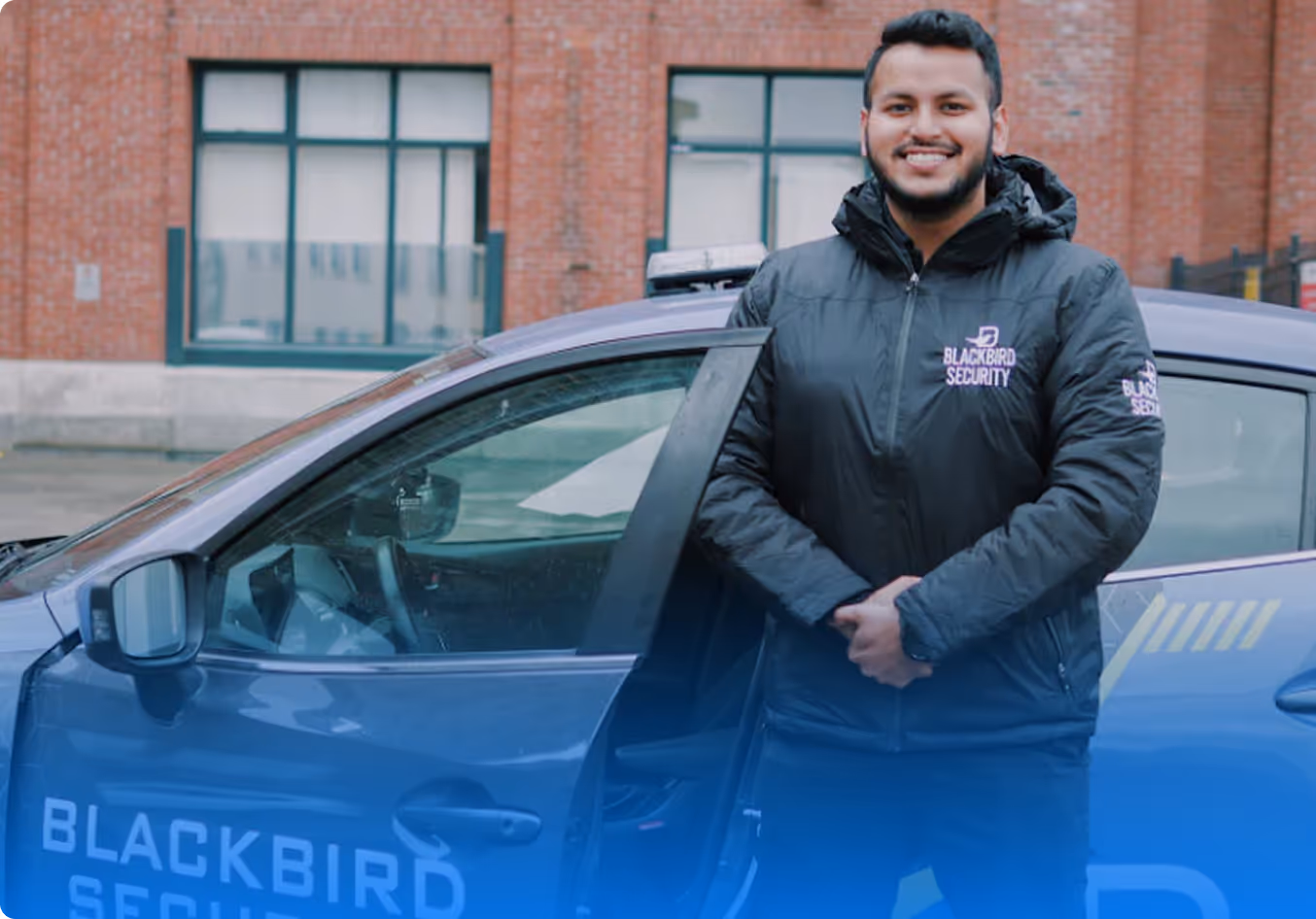 Smiling security guard standing beside an open door of a Blackbird Security vehicle in front of a brick building.