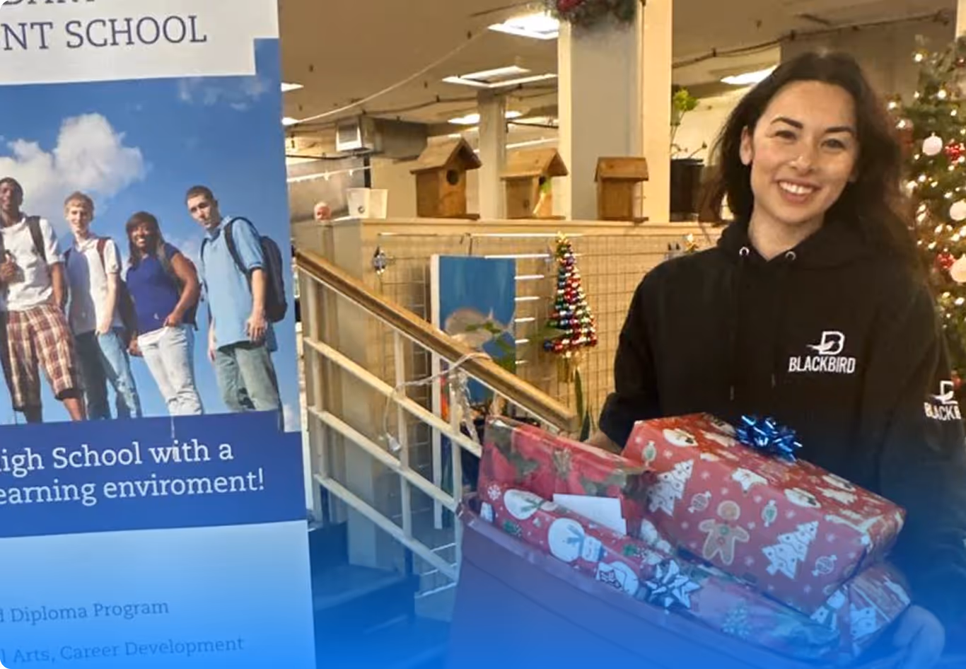 Smiling woman holding wrapped holiday gifts, standing indoors near a decorated Christmas tree and a school promotional banner.