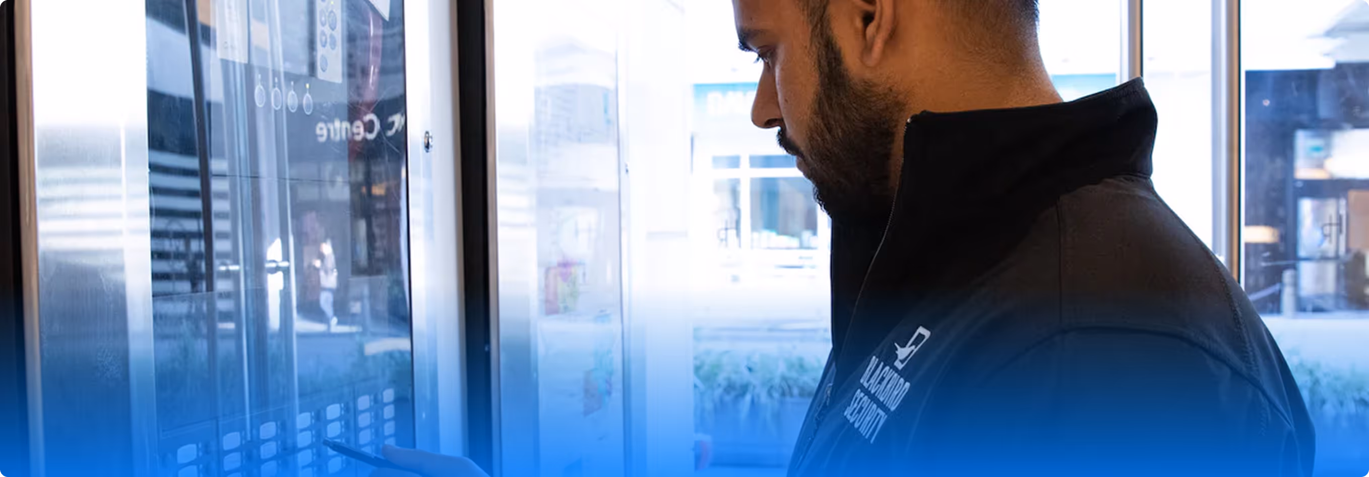 Man in a black jacket using a touchscreen kiosk or control panel indoors near a window.