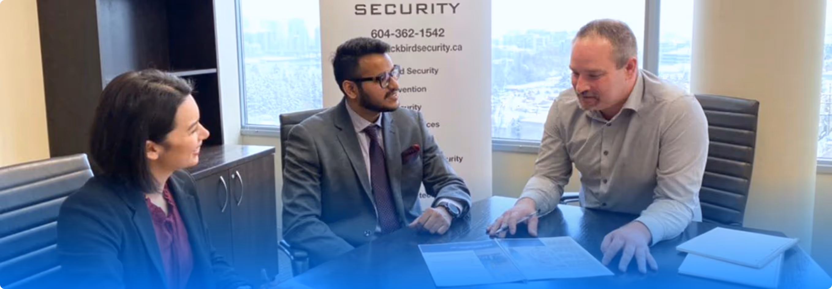 Three business professionals seated around a conference table engaged in a discussion with documents and notebooks in an office with large windows.