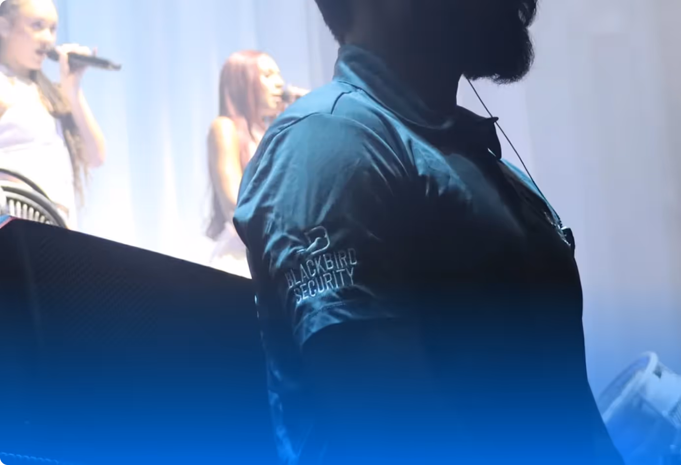 Security guard wearing a Blackbird Security shirt stands in front of two women singing on stage.