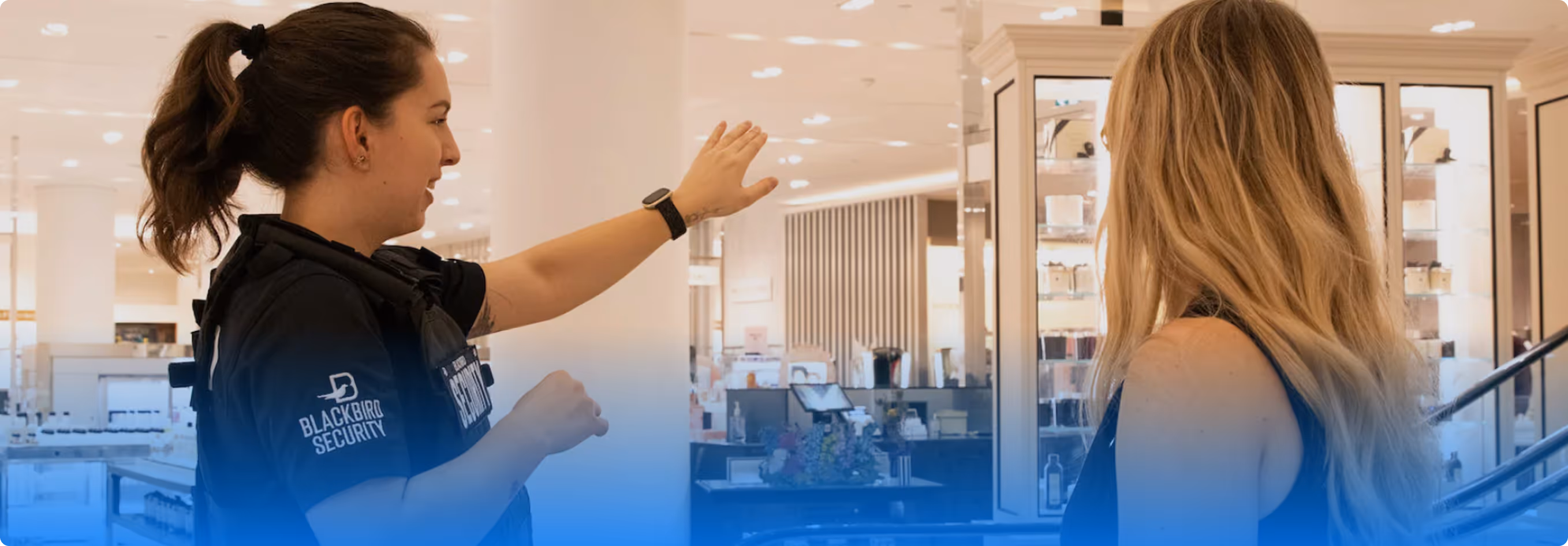 Female security guard gesturing and talking to a woman with long hair inside a bright retail store.