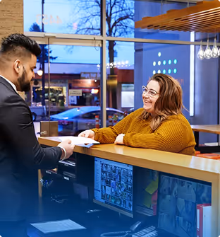 Receptionist in a mustard sweater handing a document to a man in a suit at a modern office front desk with large windows.
