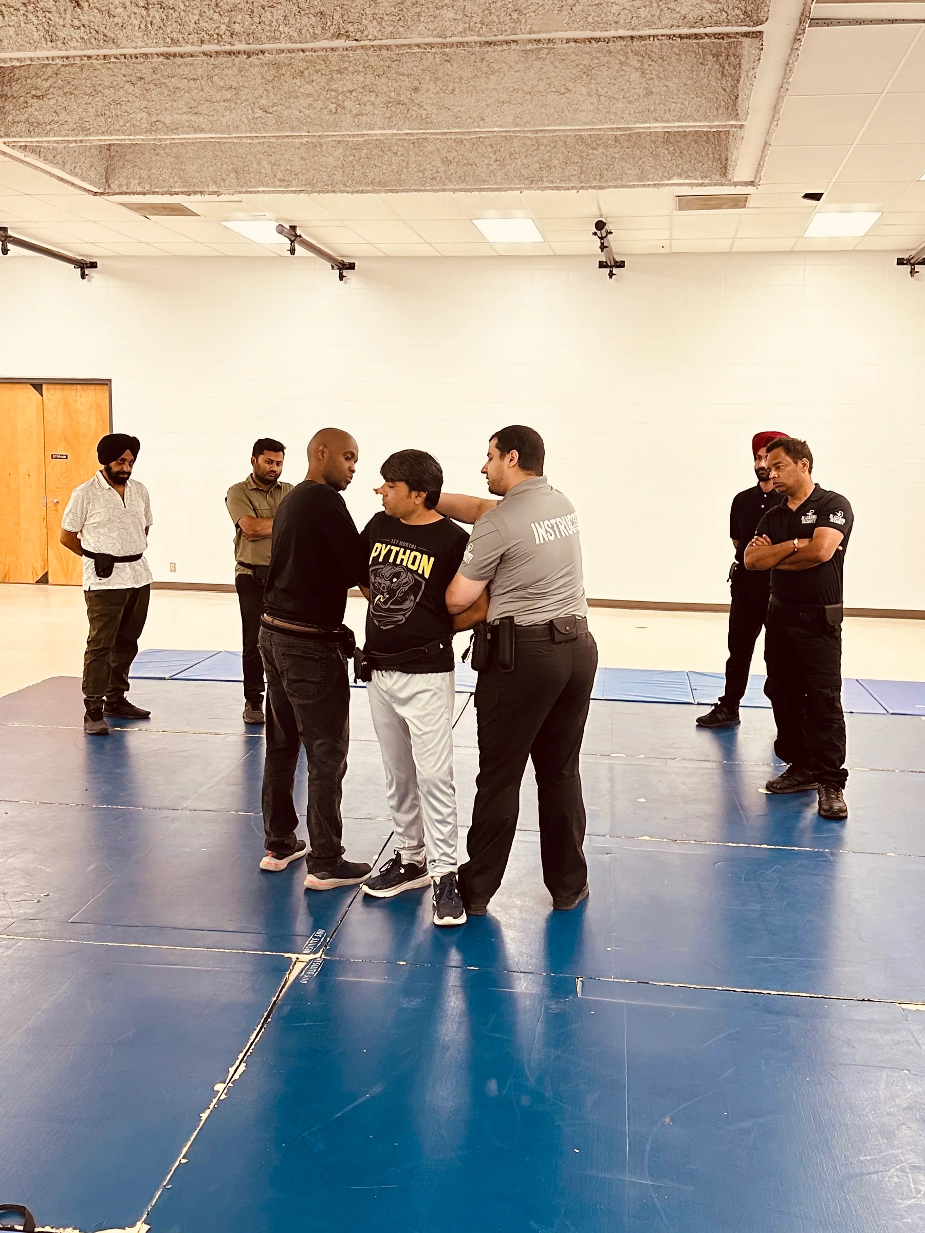 Group of men in a training room practicing a self-defense or restraint technique on a padded floor.
