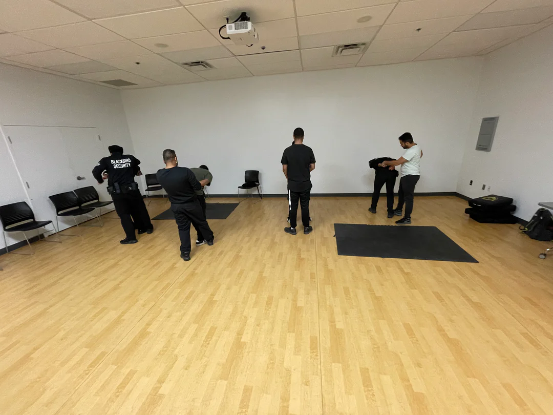 Five men in a room practicing security or self-defense techniques, with some holding others in controlled positions.