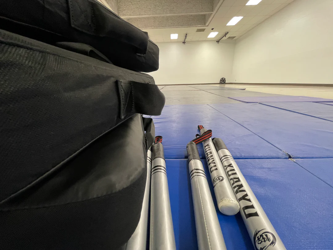 Silver martial arts sticks with red and black handles lying on blue mats next to a black padded bag in a spacious training room.