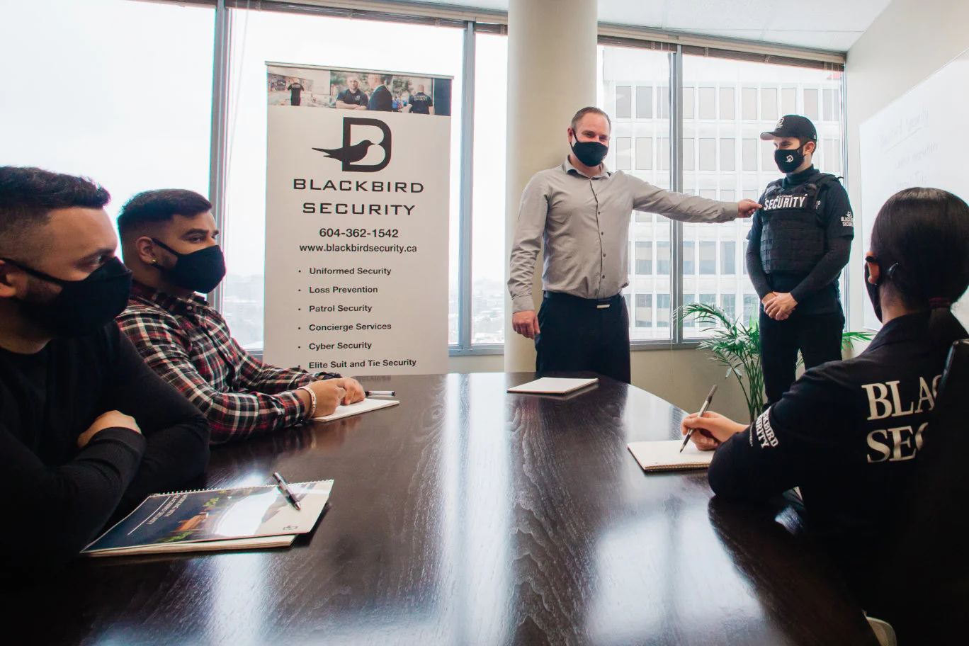 A group of people in a conference room at Blackbird Security; a man in a grey shirt gestures toward a uniformed security guard while others take notes.