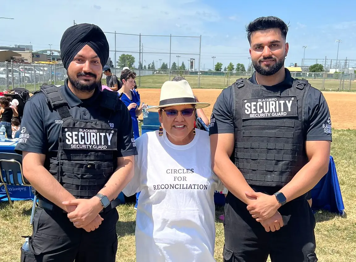 Two male security guards wearing Blackbird Security uniforms stand on either side of a smiling woman in a white 'Circles for Reconciliation' t-shirt and hat, outdoors on a sunny day.