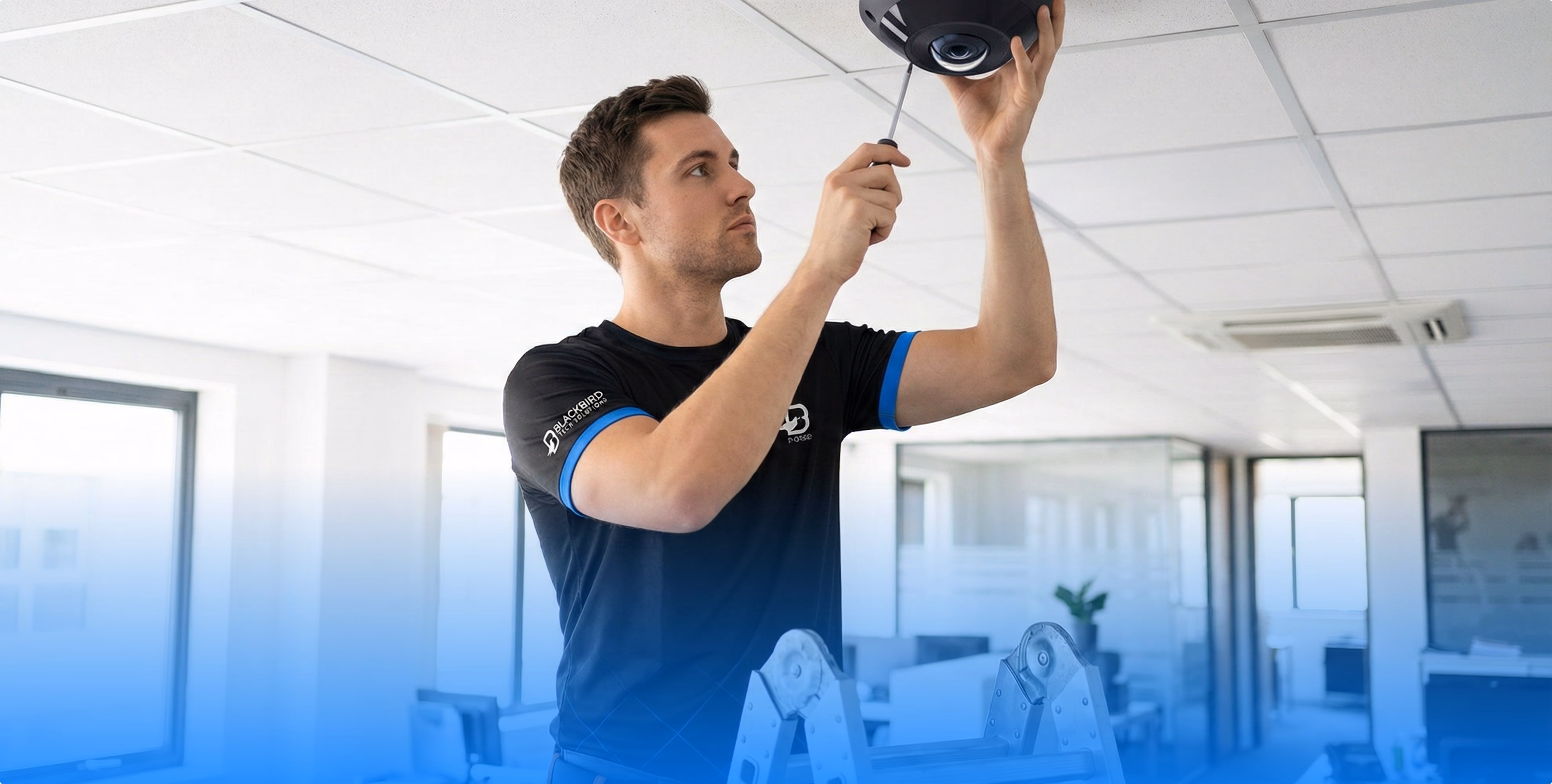 Technician wearing a black shirt with blue trim installs a ceiling security camera in a bright office setting.