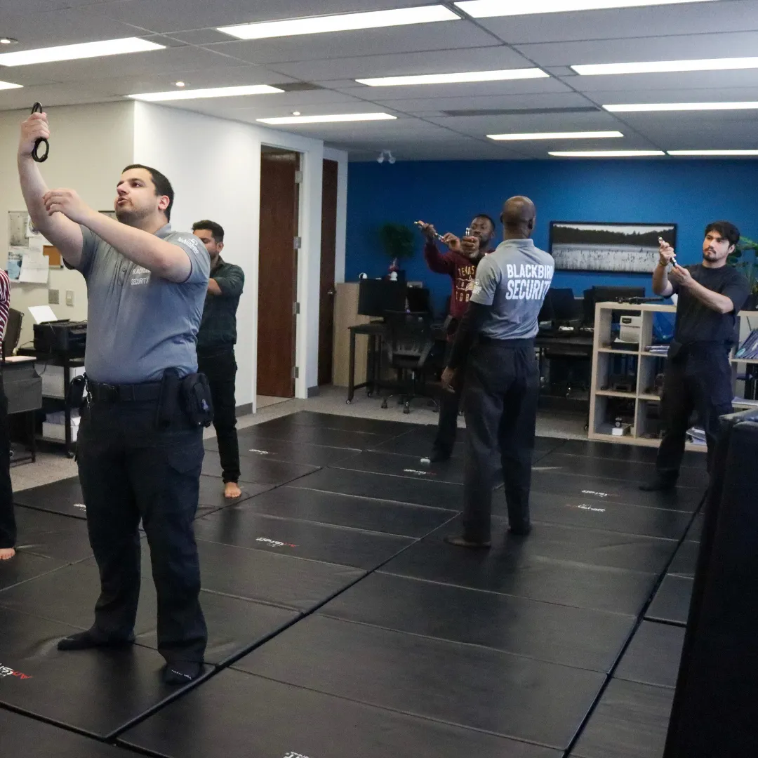 Security trainees practicing firearm handling indoors on black mats in a training room.