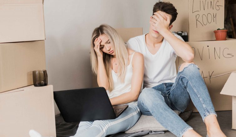Stressed couple with laptop surrounded by labeled moving boxes during relocation