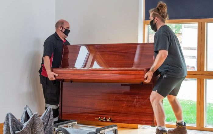 Two movers carefully lifting upright piano for safe relocation in home