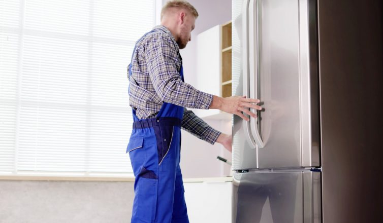 Professional mover in work uniform positioning refrigerator during appliance relocation