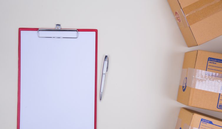 Blank clipboard with pen next to moving boxes for tracking inventory checklist