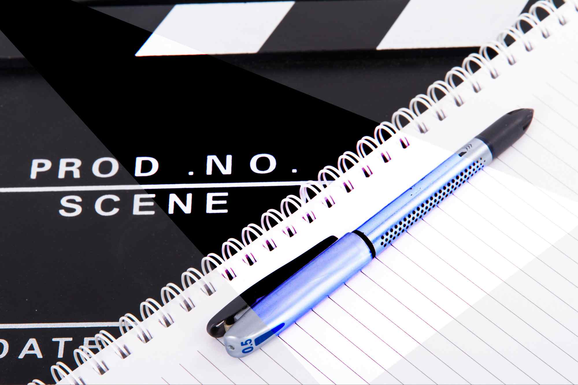Close-up of a blue pen on a spiral notebook overlaying a black film clapperboard with 'SCENE' visible, representing scriptwriting and film production planning