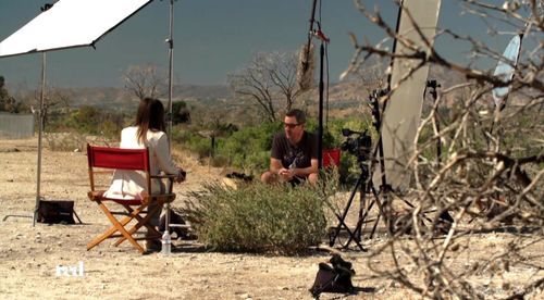Behind-the-scenes film production set in a desert landscape featuring an interview between a woman in a white blazer and a man, with professional cameras and lighting equipment visible