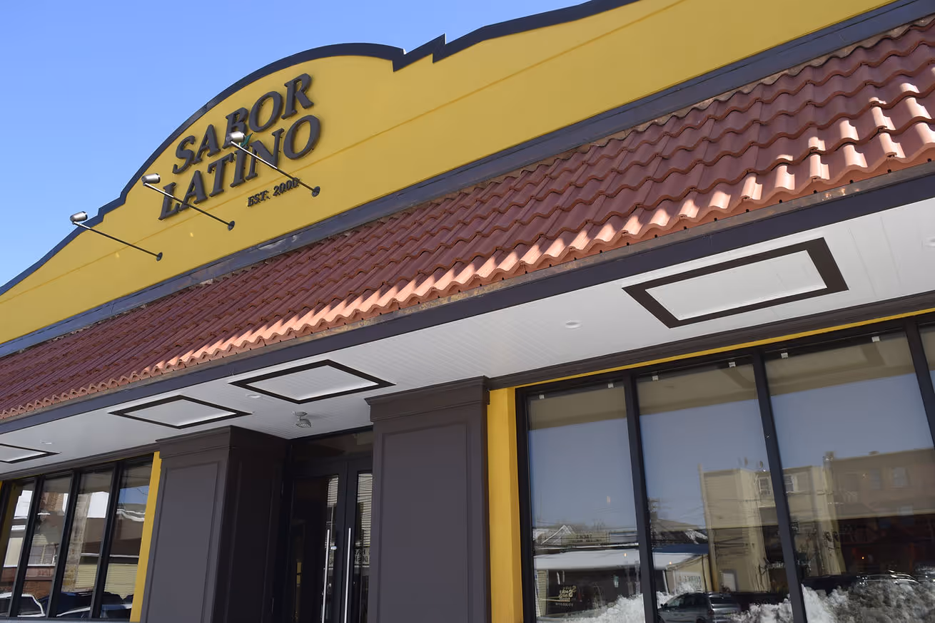 Yellow building exterior of "Sabor Latino" with red tile roof and large windows reflecting a clear blue sky. Inviting and bright tone.