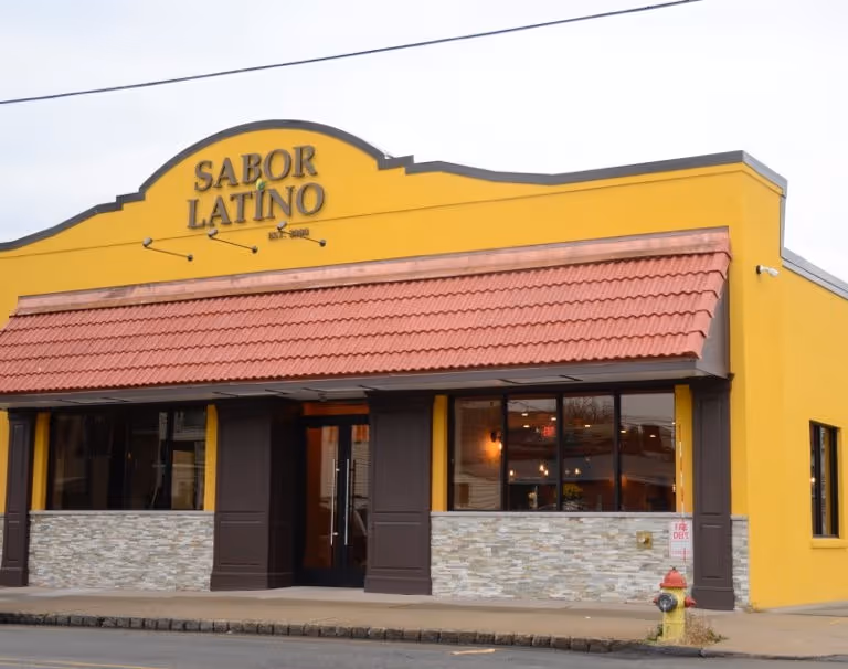 Yellow building with a red-tiled roof labeled "Sabor Latino" on top. Large windows reveal a warmly lit interior. Sidewalk and fire hydrant in front.