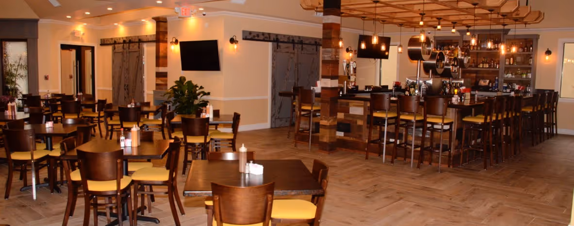 Spacious, warmly lit restaurant interior with wooden chairs and tables. A large bar with high stools lines the right side.