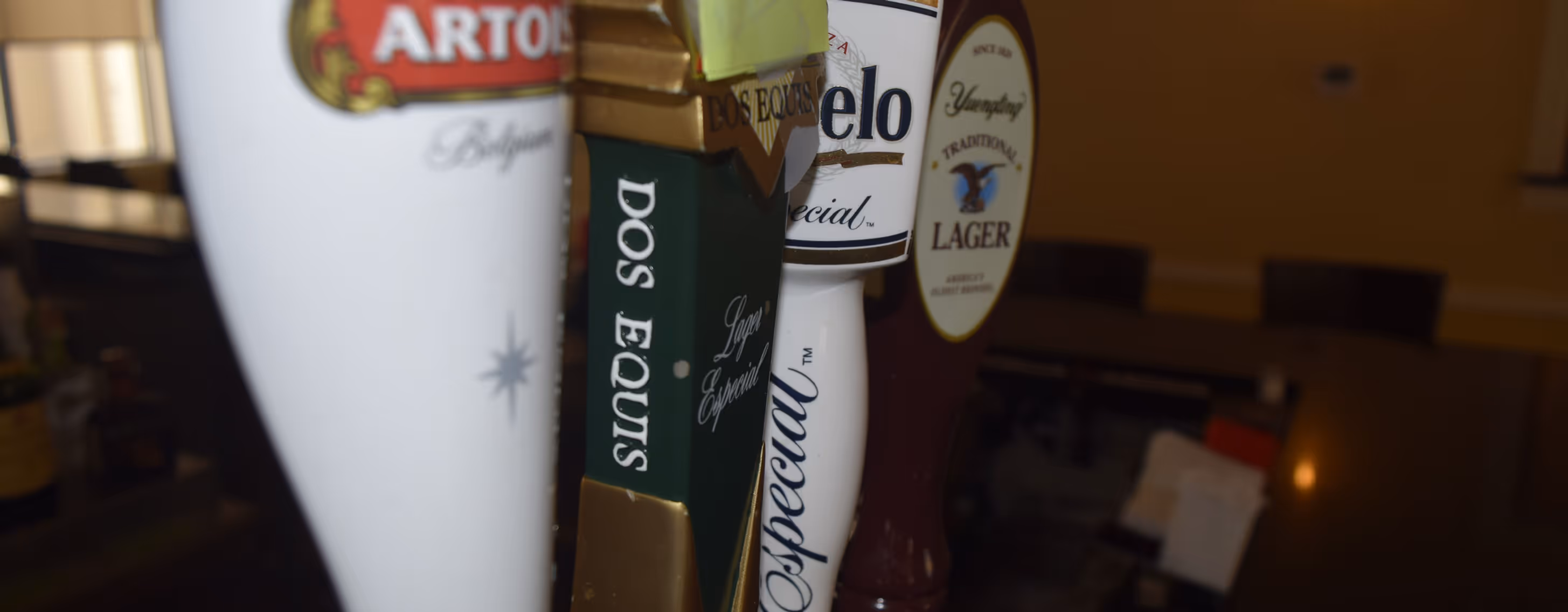 Close-up of beer tap handles in a dimly lit bar, featuring brands like Dos Equis and Modelo.