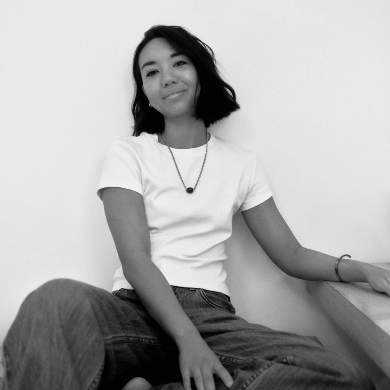 Miranda with shoulder-length hair smiling, wearing a white t-shirt and jeans, sitting casually against a plain wall.