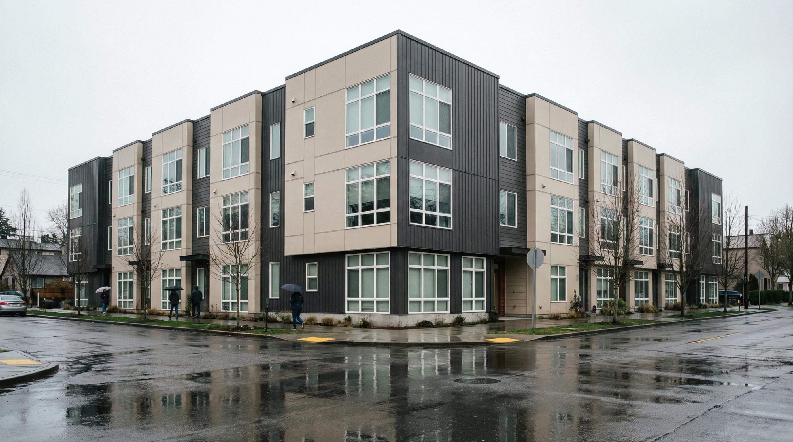 Modern three-story apartment building on a rainy day with wet streets and people walking under umbrellas.