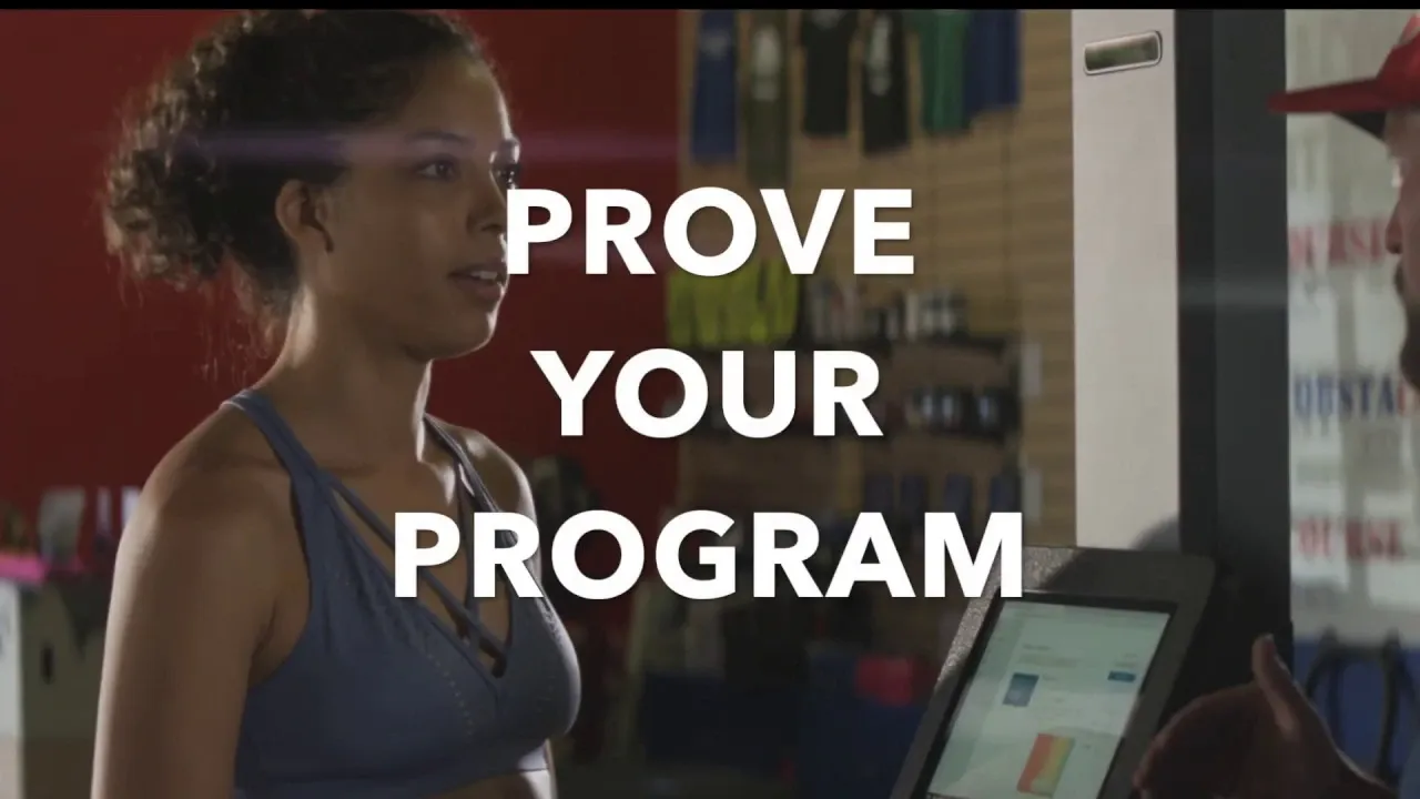 A woman wearing a blue tank top and a sports bra stands in front of a treadmill monitor.