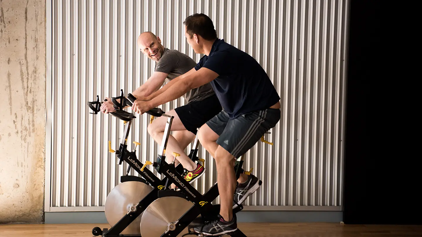 Two men are on exercise bikes in front of a wall.