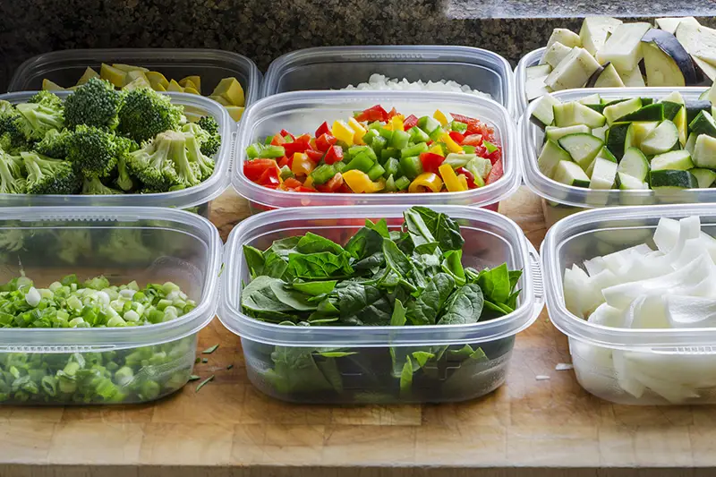 A wooden cutting board with a variety of vegetables in plastic containers.