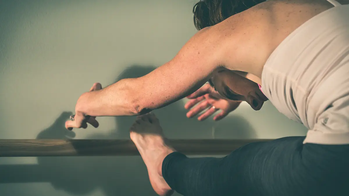 Woman stretching her arms forward while sitting with one leg extended on a ballet barre.