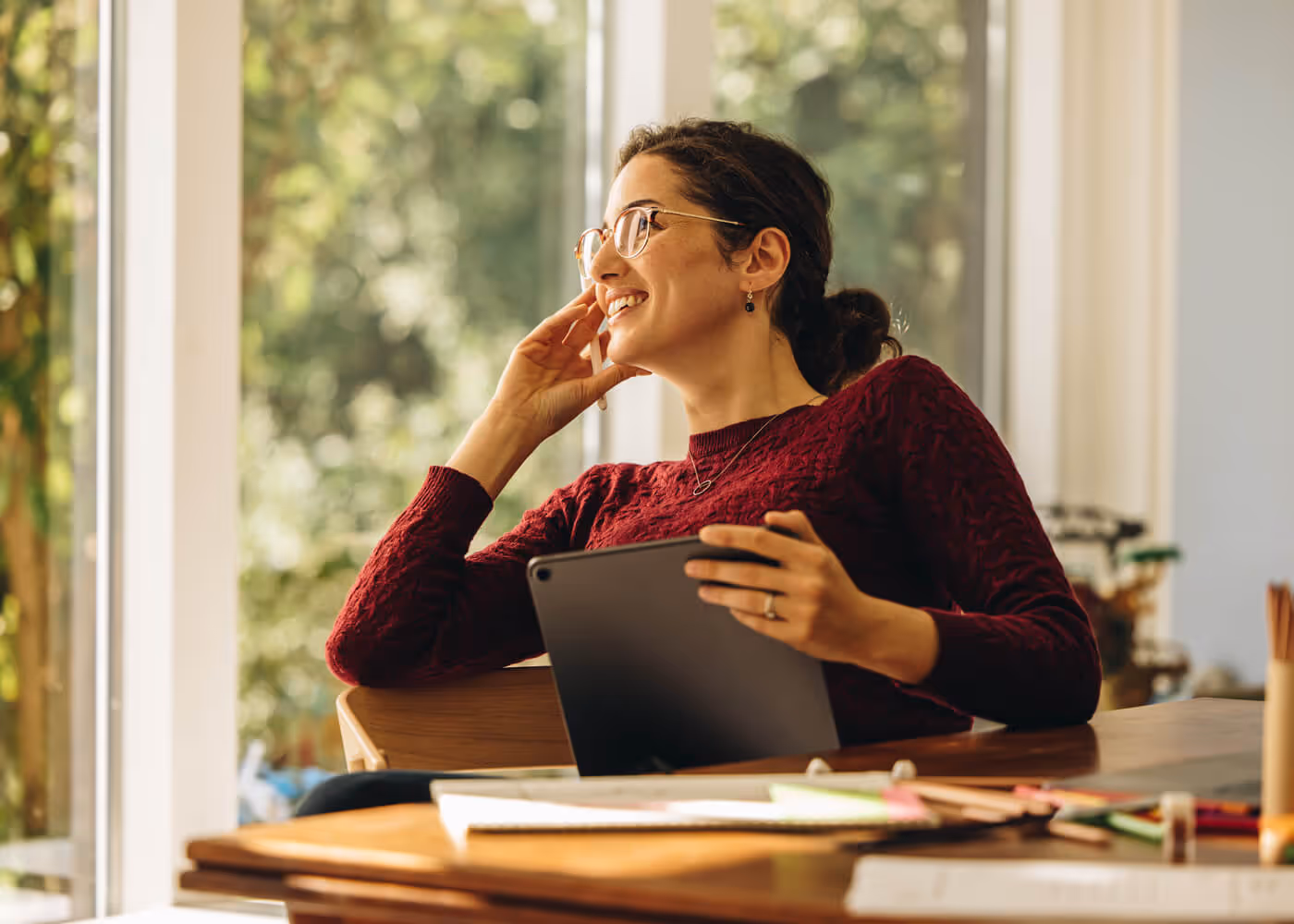 Smiling woman wearing glasses talks on a phone while holding a tablet at a table near a bright window.