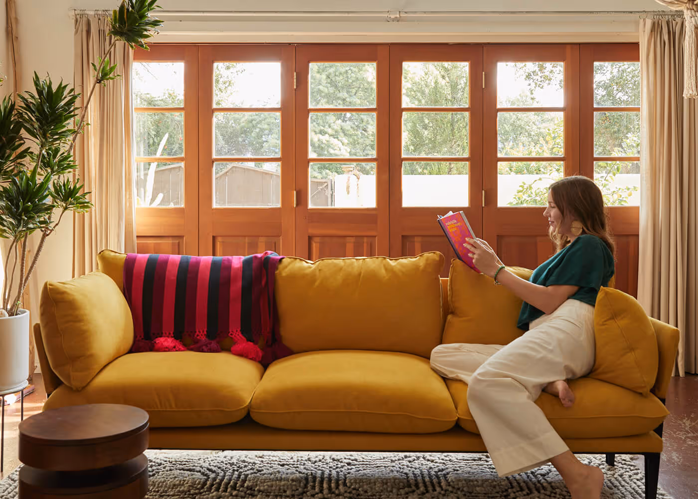 Woman relaxes on a mustard-yellow sofa reading a book in a bright living room with large windows and plants.