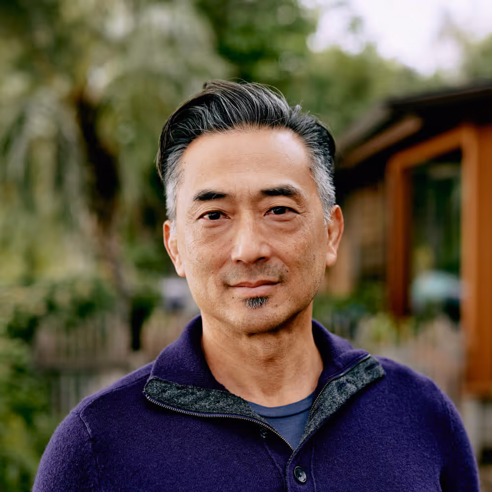Portrait of a middle-aged man outdoors, standing in front of a home and trees with a calm expression.