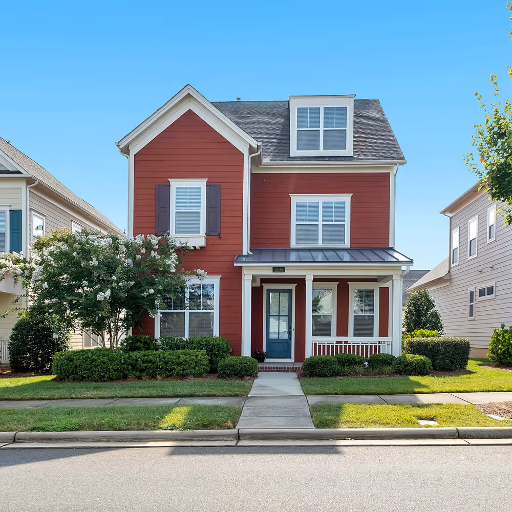 Red two-story suburban home with a front porch, manicured shrubs, and a walkway leading to the front door.