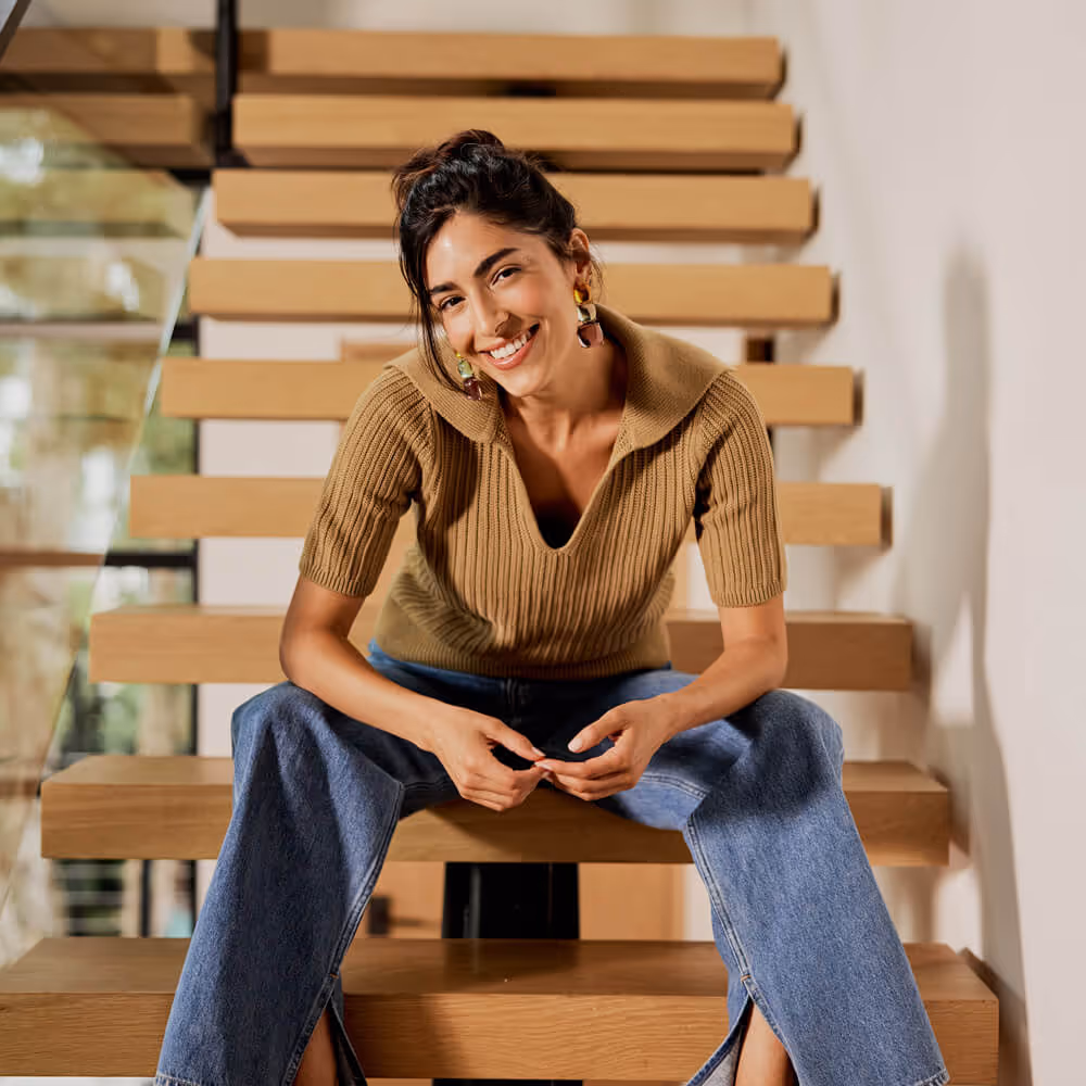 Smiling woman sits on modern wooden stairs indoors, leaning forward with hands clasped.