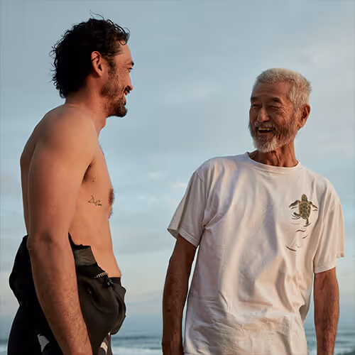 Two men laugh and talk together at the beach under an open sky.