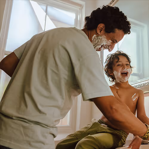 Father and young child laugh during a playful shaving routine in a bathroom, with shaving cream on their faces.