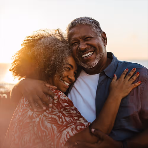 Smiling couple embraces at the beach during sunset with warm sunlight in the background.