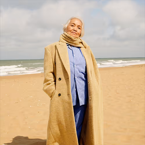 Older woman stands on a windy beach wearing a long coat and scarf near the shoreline.