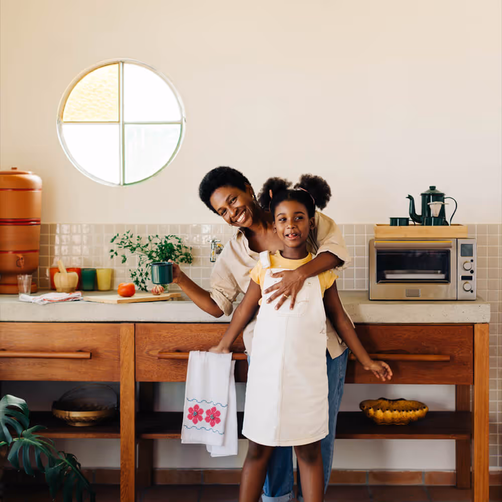Mother hugs her daughter in a bright kitchen while holding a mug near the countertop.