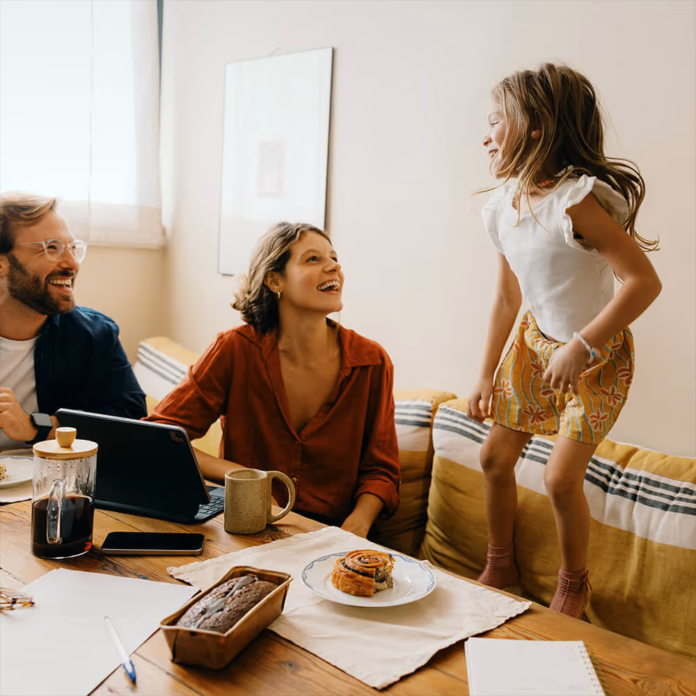 Family smiles together at home as a young girl dances on the couch beside her parents, with a coffee table and tablet in front.