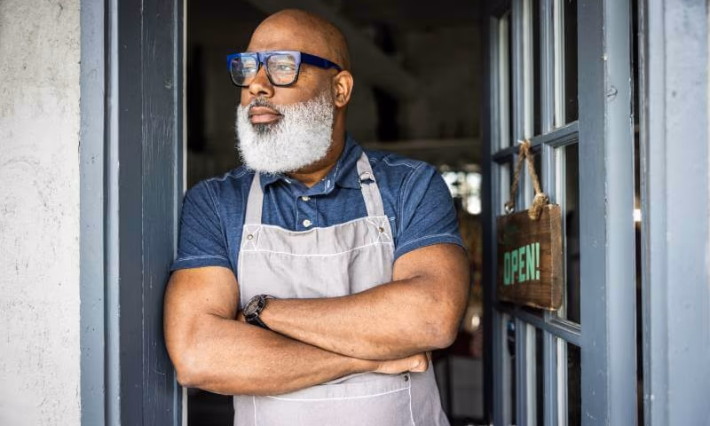 Smiling small business owner in an apron stands with arms crossed at his shop doorway.